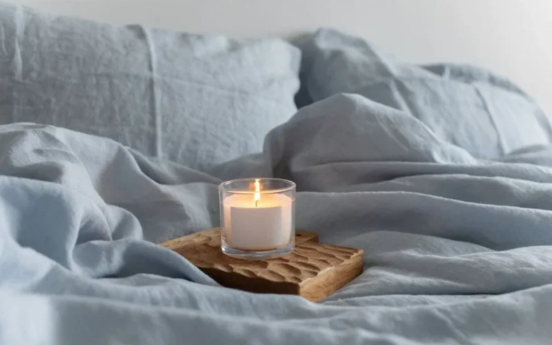 A soothing scene with a lighted candle on a wooden tray amidst soft blue linen bedding.