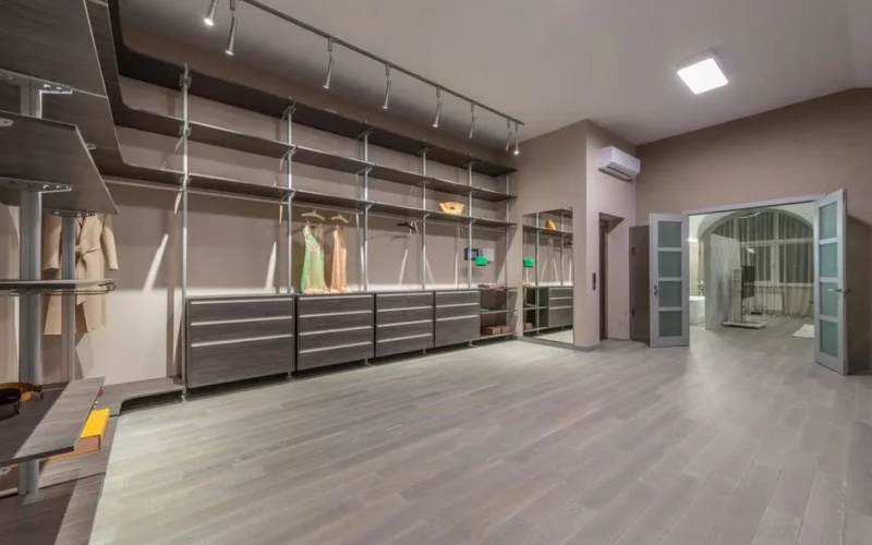 Stylish dresses and coat hanging on hangers in spacious walk in closet with empty shelves and opened doors in modern apartment