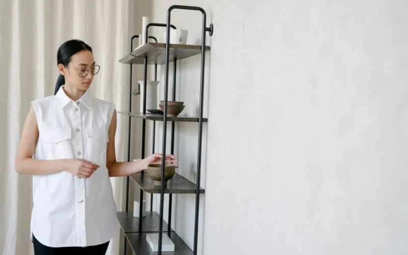 A woman in a white shirt arranges minimalist home decor on an industrial-style shelving unit.