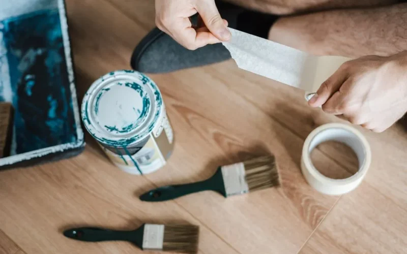 From above of crop anonymous male unwrapping sticky tape roll on parquet near paint brushes in house