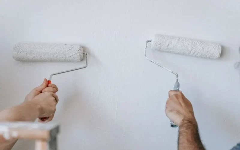 Close-up of two individuals painting a wall with paint rollers indoors.