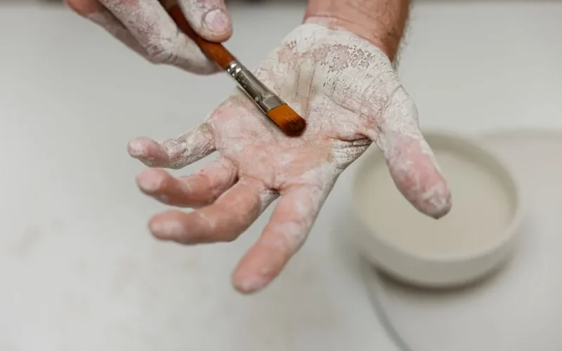 Artisan's hands covered in clay, using a brush in a creative workspace.