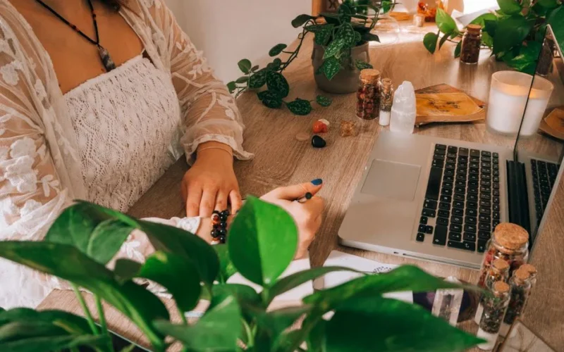 A peaceful workspace featuring a woman writing, surrounded by greenery, crystals, and a laptop.