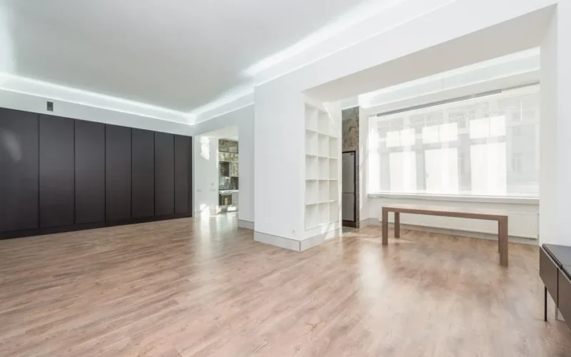 A modern minimalist room interior with hardwood flooring and sunlight streaming in through large windows.