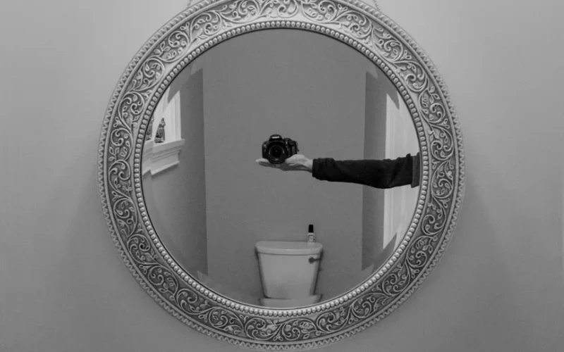 Intricate round mirror capturing a camera reflection in a monochrome bathroom setting.