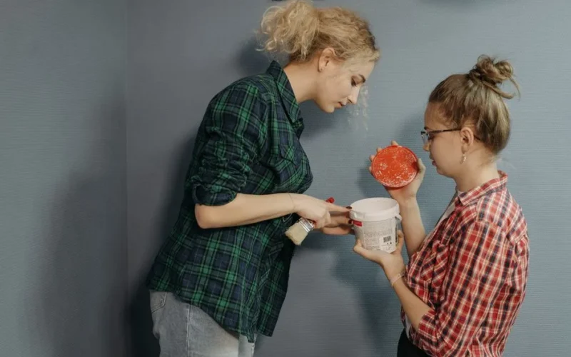 Two women collaborating on painting a wall, holding paint and brushes, in a modern setting.