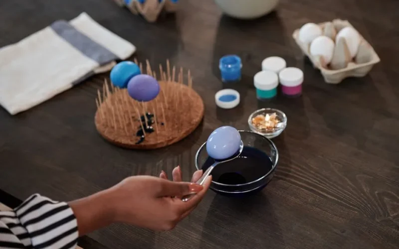 Hand dipping an egg into blue dye, surrounded by painting supplies, for Easter preparations.