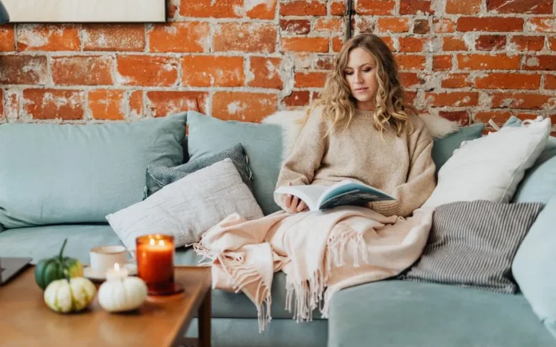 A woman enjoys a peaceful reading moment on a comfy sofa with lighted candles and a warm, cozy ambiance.