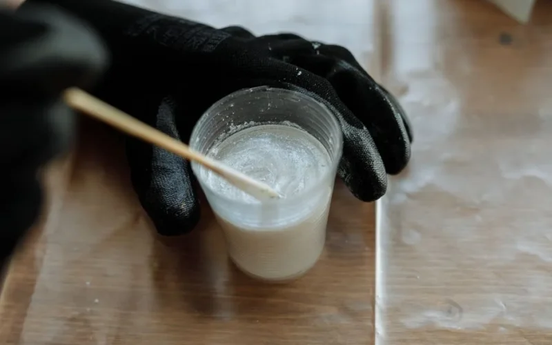 Detailed view of hands with gloves mixing resin in a plastic cup with a stirrer on a wooden surface.