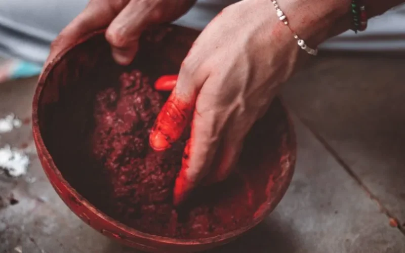 Close-up of a hand mixing vibrant red pigment powder in a clay bowl.