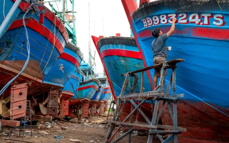 A man painting hulls of vibrant boats in an outdoor shipyard.