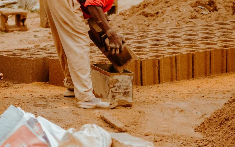 A worker shaping bricks with a concrete mold at an outdoor construction site.