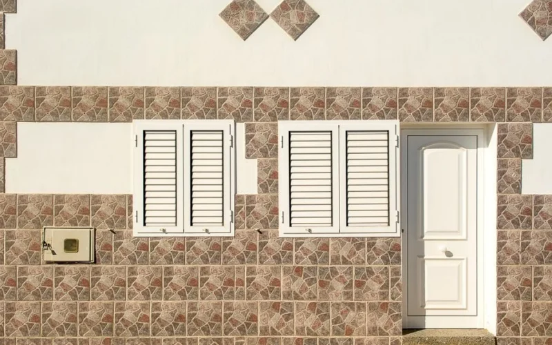 Close-up of a building facade featuring two white shuttered windows and a white door set within a mosaic tile wall.