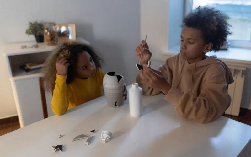 Two children collaborate to fix a broken vase using glue on a table at home.
