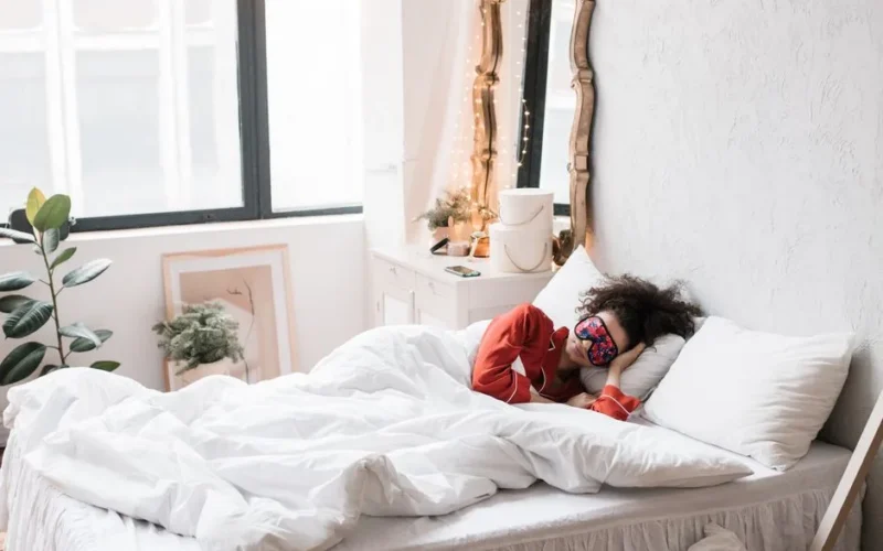 Woman wearing a sleep mask, resting on a cozy white bed surrounded by indoor plants in a bright bedroom.
