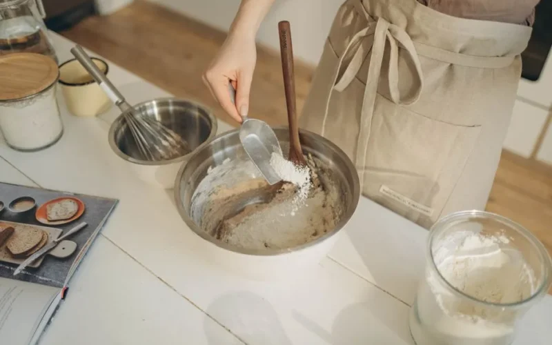 Person adding flour to dough in a stainless steel bowl while baking at home.