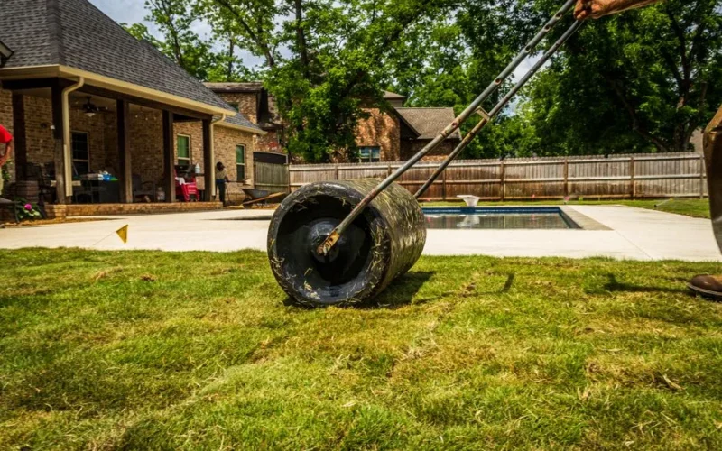 A worker rolling a lawn in a suburban backyard near a pool on a sunny day.