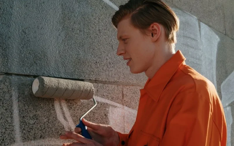 Young adult participating in community service, painting over graffiti on a concrete wall.