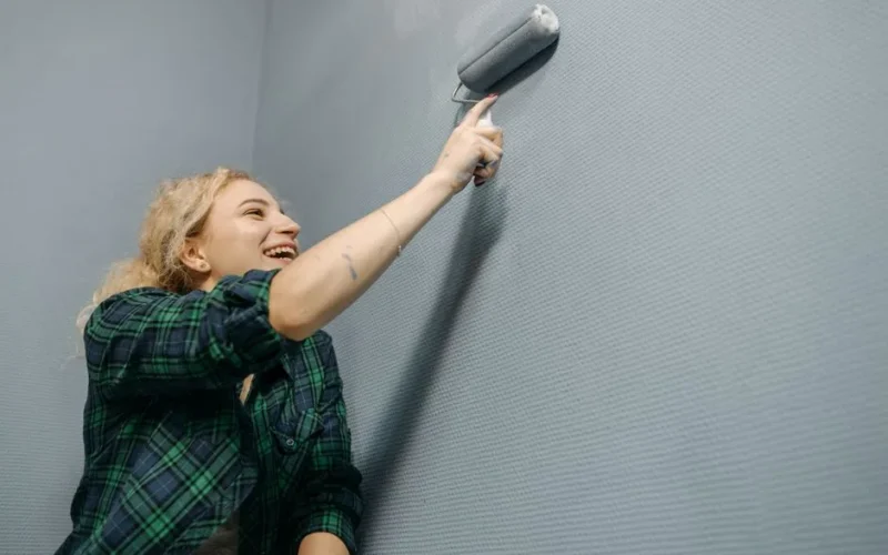 Laughing woman painting a blue wall indoors with a roller brush during home renovation.
