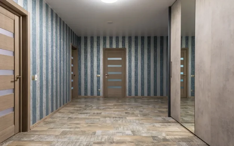 Stylish hallway with mirror on wardrobe and various wooden doors near walls with striped wallpaper in modern flat with glowing chandelier