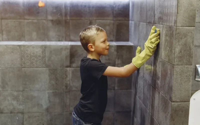 Side view of kid with blond hair in yellow rubber gloves cleaning tile wall in bathroom