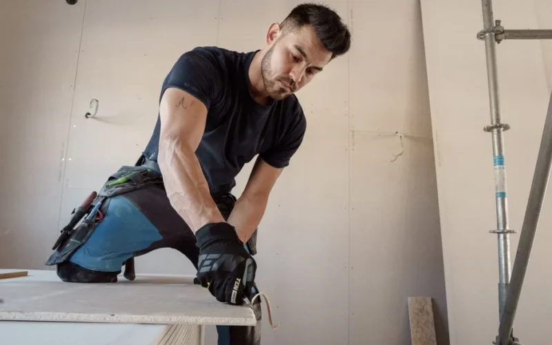 Man in gloves cutting plasterboard for drywall installation inside a building.