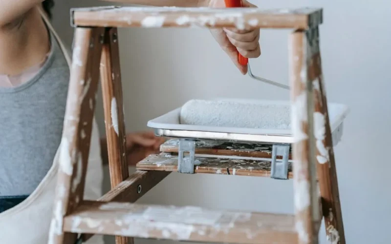 Adult woman using a roller and ladder to paint a room wall indoors.