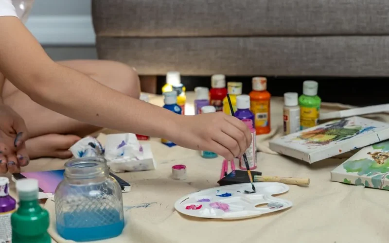 A close-up of a hand painting with acrylics in a cozy indoor setting, showcasing colorful art materials.