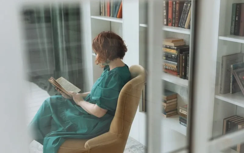 A woman enjoys reading in a comfortable chair surrounded by books in a home library.