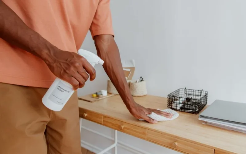 A person cleaning a wooden desk with a spray bottle and cloth, showcasing tidiness.