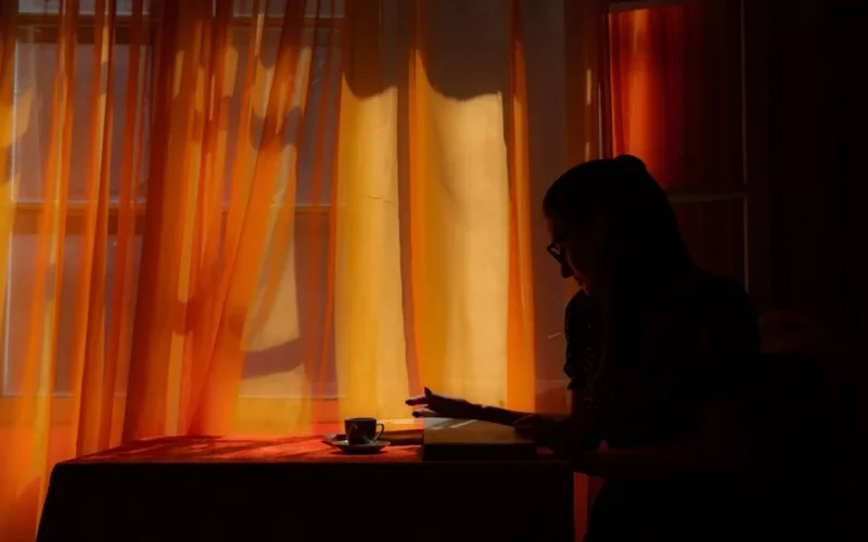 A silhouette of a woman reading a book near a window with golden curtains and soft light.