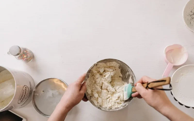 A person mixing homemade dough in a bowl with baking utensils on a white table.