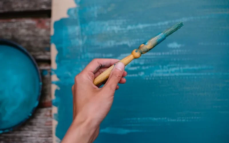 Top view of anonymous person using paintbrush to color carton in azure paint placed on wooden bench near bowl with gouache