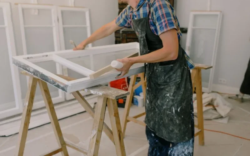 Unrecognizable male master wearing dirty apron standing near window with white frames on wooden workbench while working in professional studio