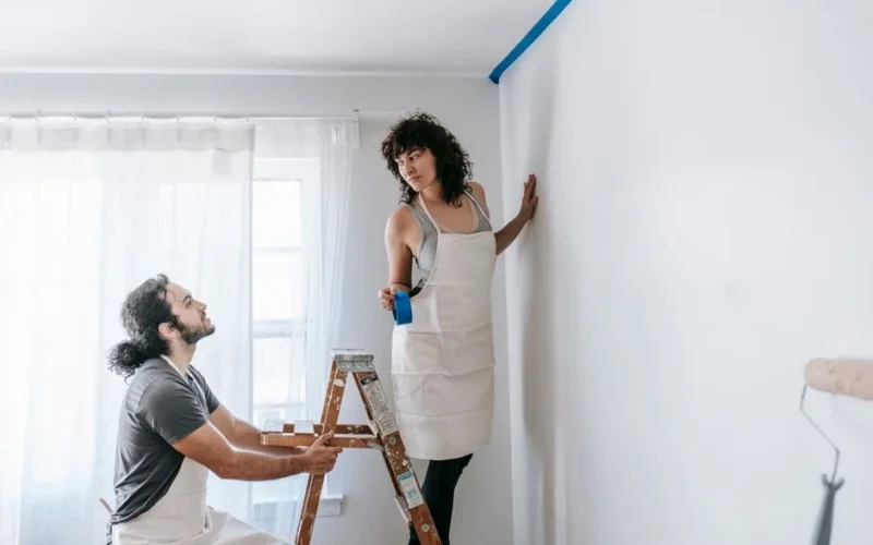 A couple in aprons working together on room renovation using paint rollers indoors.