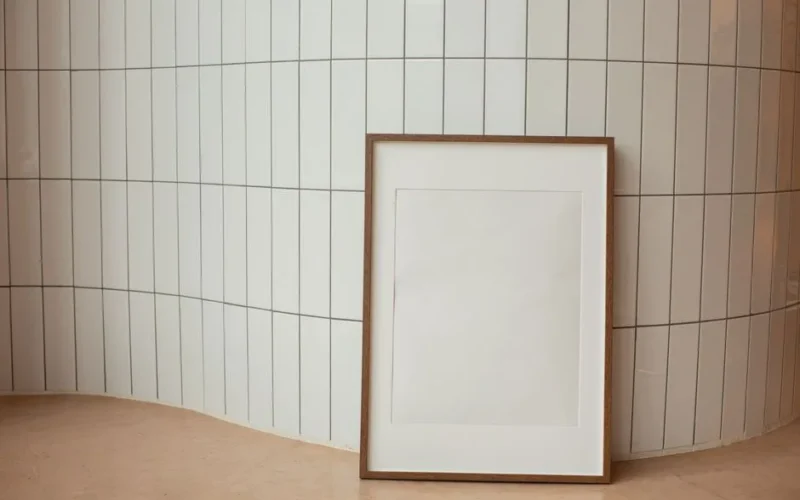 Empty white wooden photo frame placed on floor against curvy tiled wall in light room