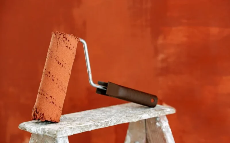 Detailed image of a paint roller resting on a ladder with a vibrant orange wall in the background.