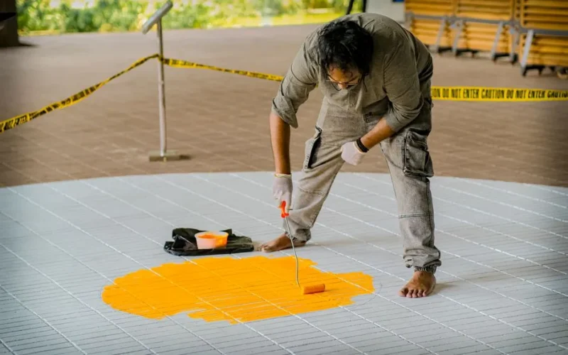 An artist paints a bright yellow circle on tiles in a creative outdoor project.