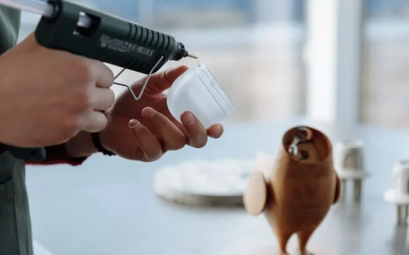 Close-up of hands using a glue gun to craft a wooden figurine in a workshop setting.