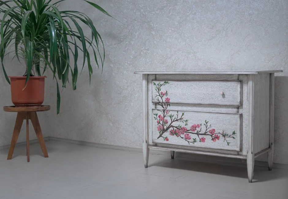 A vintage cabinet with floral design next to an indoor plant on a wooden stand.