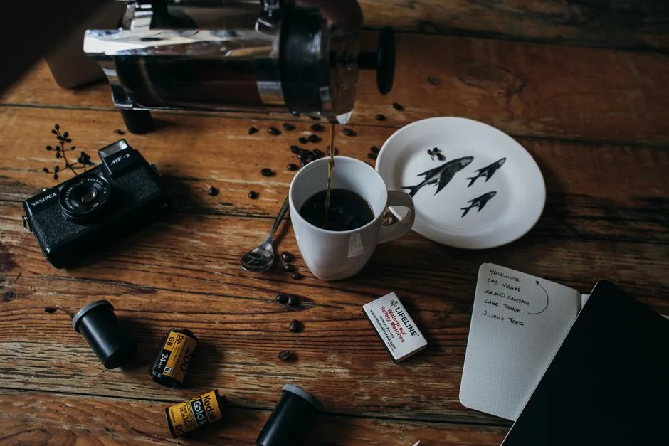 From above freshly brewed coffee pouring into cup placed on messy table with old photo camera and films