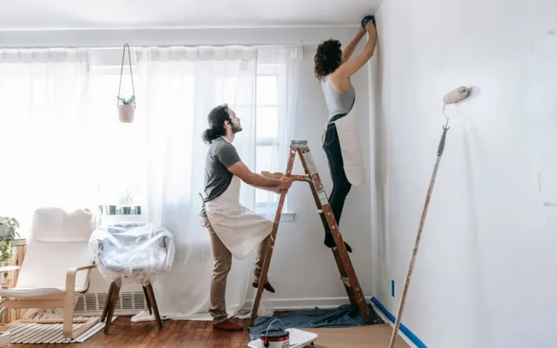Couple renovating their home, with man supporting ladder and woman working on walls indoors.