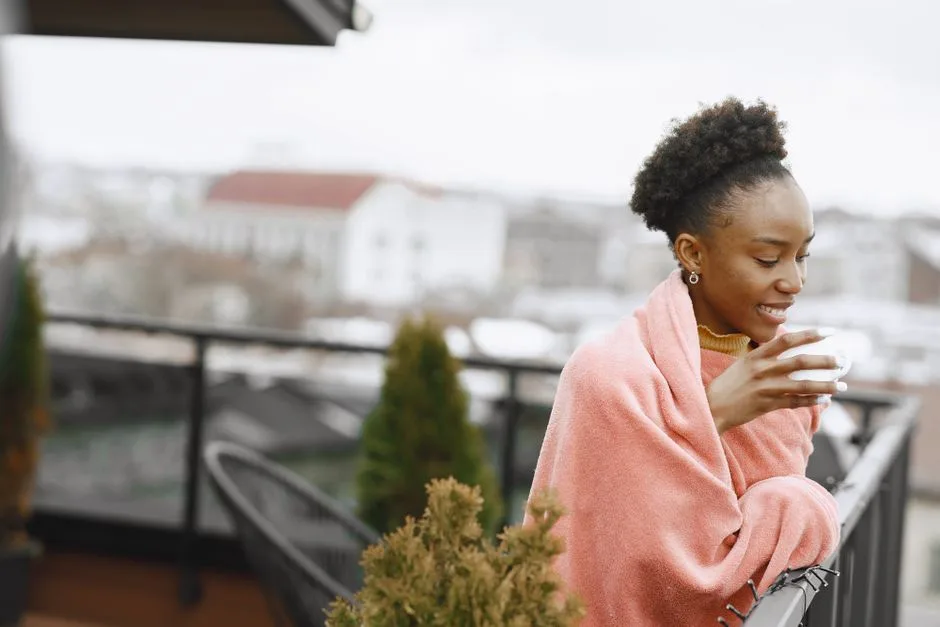 Woman enjoying a hot drink on a balcony wrapped in a pink blanket.